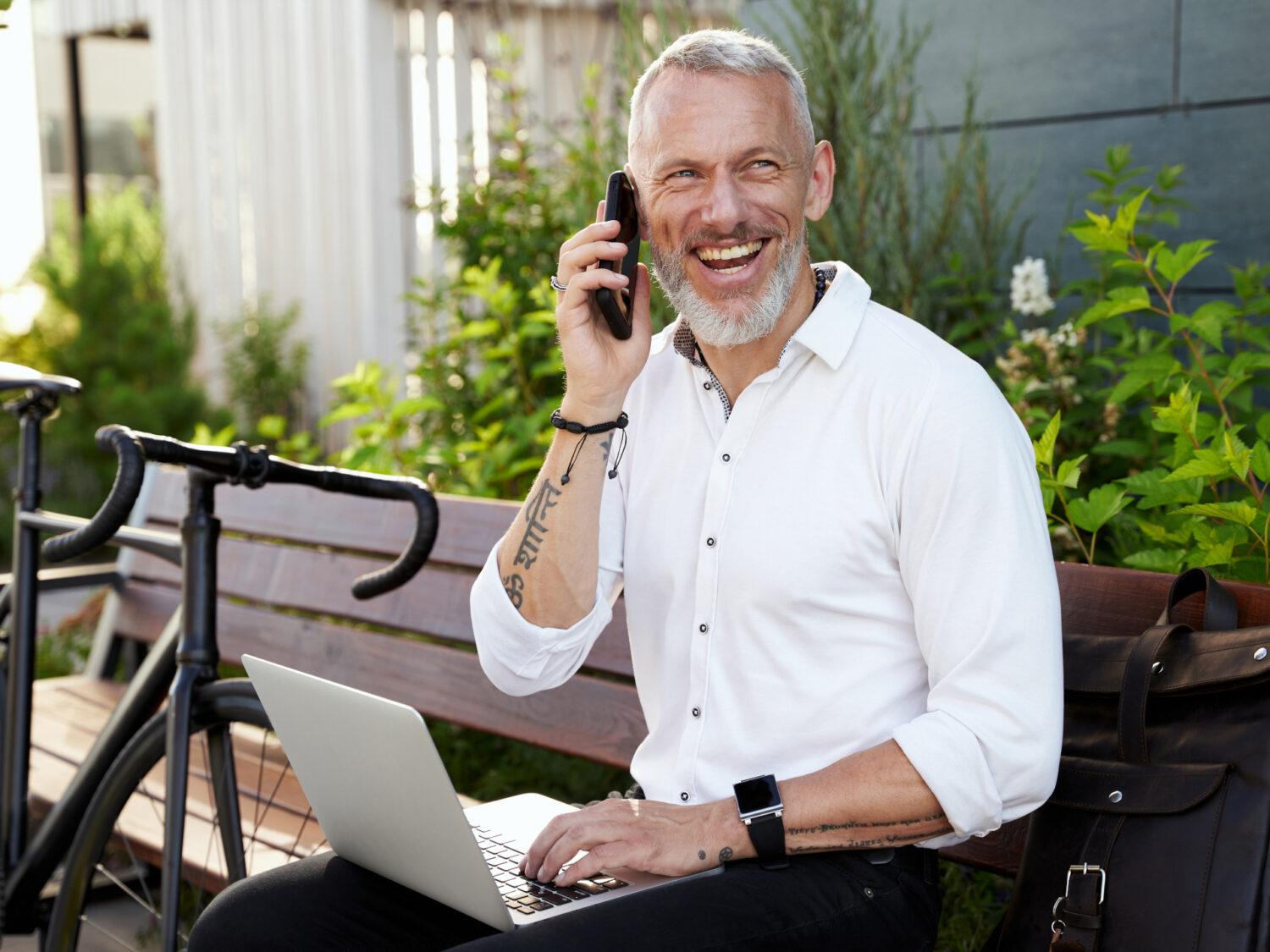 Cheerful middle aged businessman smiling, talking on the phone, using his laptop while sitting on the bench outdoors with a bicycle next to him. Business, career concept