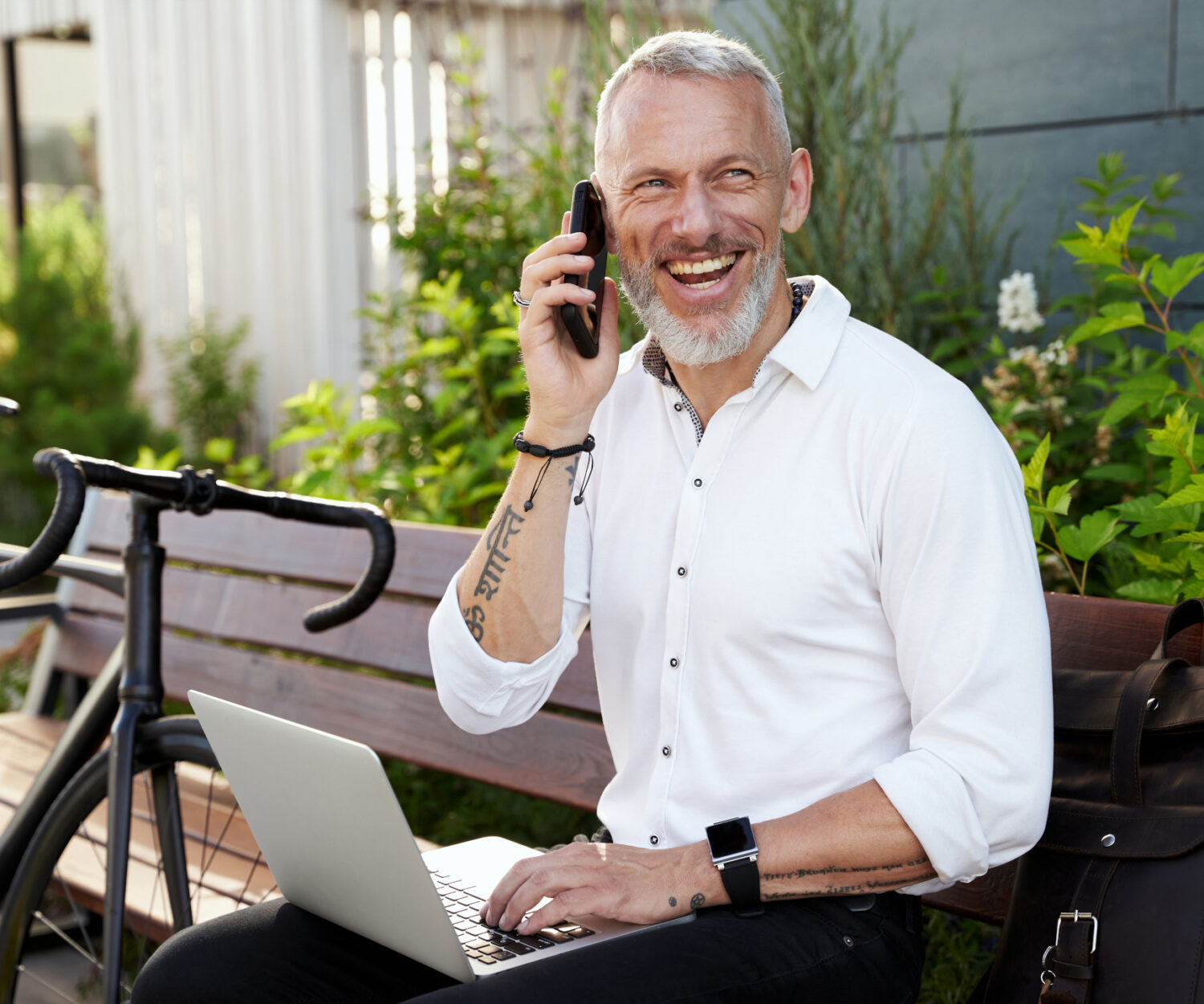 Cheerful middle aged businessman smiling, talking on the phone, using his laptop while sitting on the bench outdoors with a bicycle next to him. Business, career concept